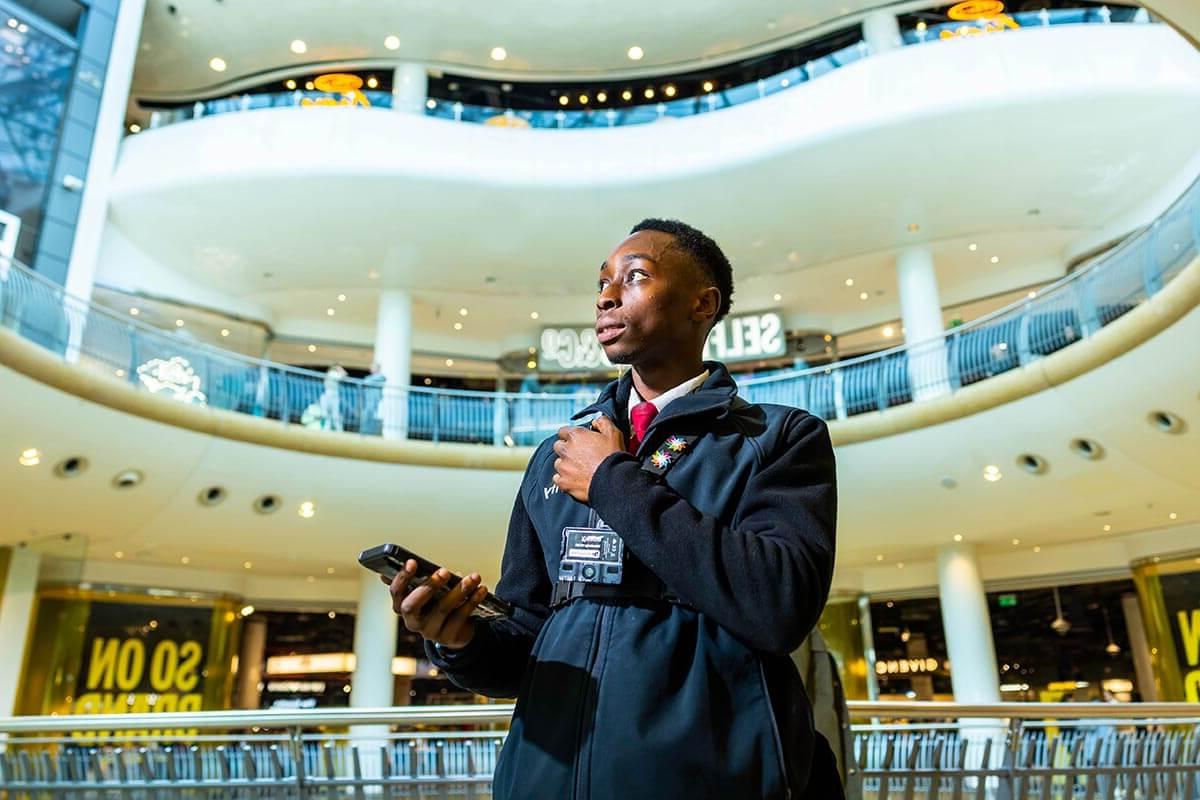 A young black 安全 guard holding his radio, with levels of a shopping centre seen behind him